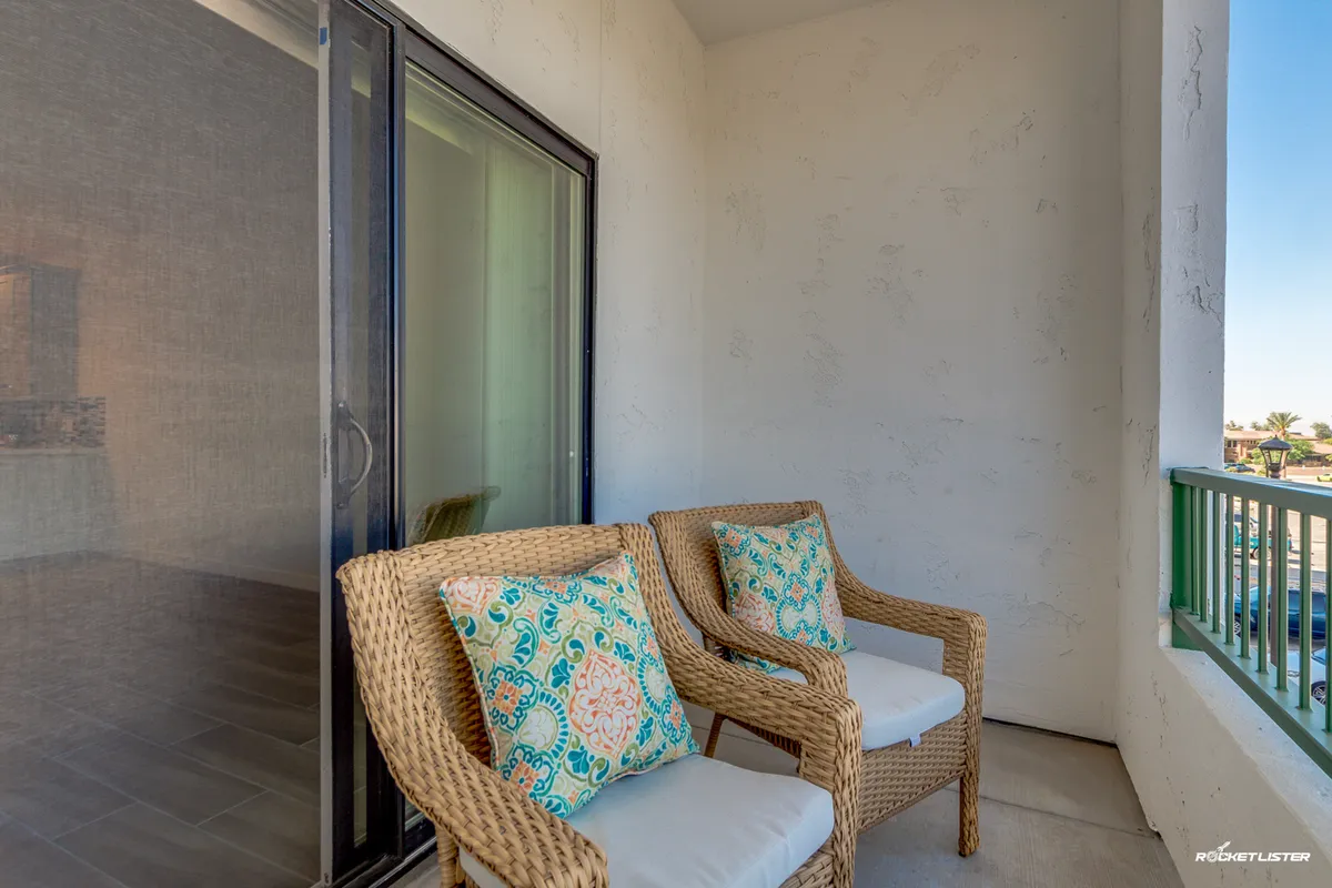 A cozy private balcony setup featuring two wicker chairs with colorful cushions facing outwards. The balcony is accessible through a sliding glass door, and the setting suggests a relaxing space to enjoy morning coffee or unwind in the evening. The soft outdoor lighting hints at ideal sun patterns.