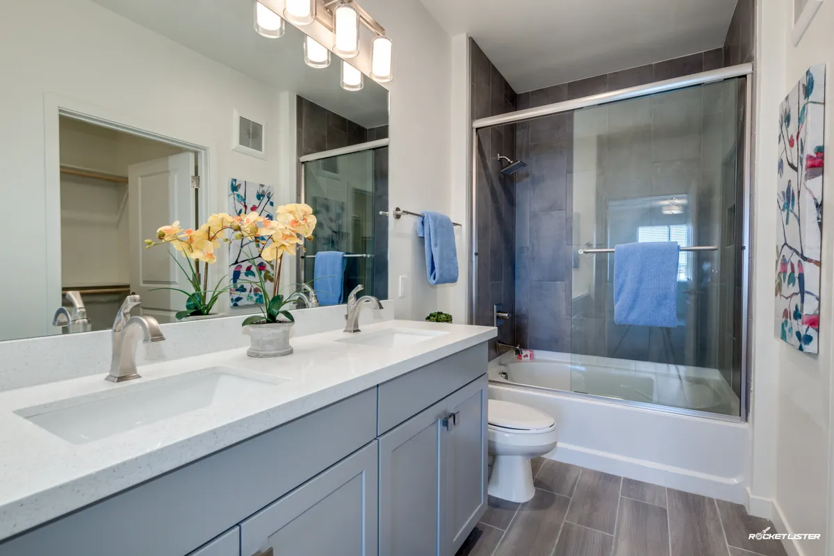 A bright, modern bathroom featuring a double sink vanity with quartz counters, a framed mirror, a glass-enclosed shower, and a decorative flower arrangement. The space feels spacious and luxurious, perfect for morning routines or unwinding at night.