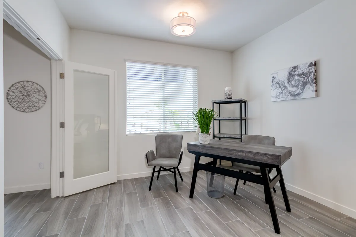A focused, well-lit office space featuring a desk, chairs, shelves, and a window bringing in natural light. The scene suggests a quiet environment ideal for working, studying, or reading, and the practical furnishings support both productivity and comfort.