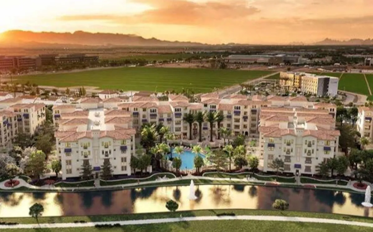 A vibrant exterior shot featuring apartment buildings with terracotta roofs surrounding a large, inviting swimming pool. Manicured lawns and a fountain enhance the resort ambiance, while the sunset sky creates a warm, relaxed mood. The scene hints at opportunities for poolside relaxation, evening strolls, and community gatherings.