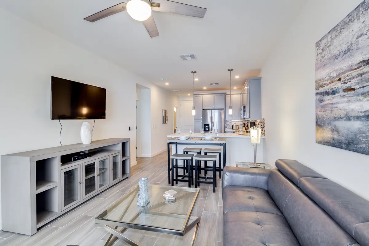 A welcoming, contemporary living area featuring a comfortable gray sofa, sleek modern coffee table, large flat-screen TV, abstract wall art, and a kitchen area with bar stools visible in the background. The space is bathed in natural light from oversized windows, and the open layout connects the living and kitchen areas seamlessly.