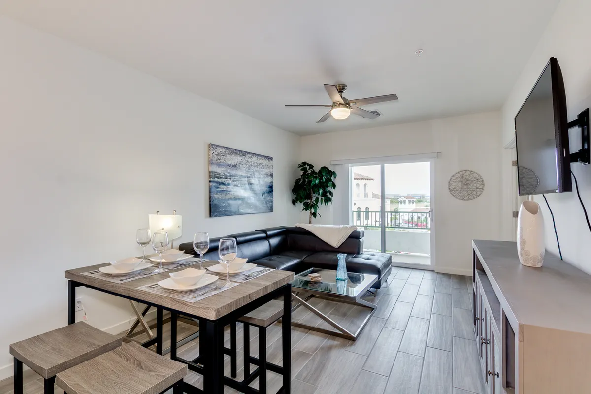 A cozy and contemporary interior living space featuring a dining table set for two, a modern black leather sofa, and a large window with a clear view outside. Natural light fills the room, hinting at a connection between home and the vibrant world beyond.