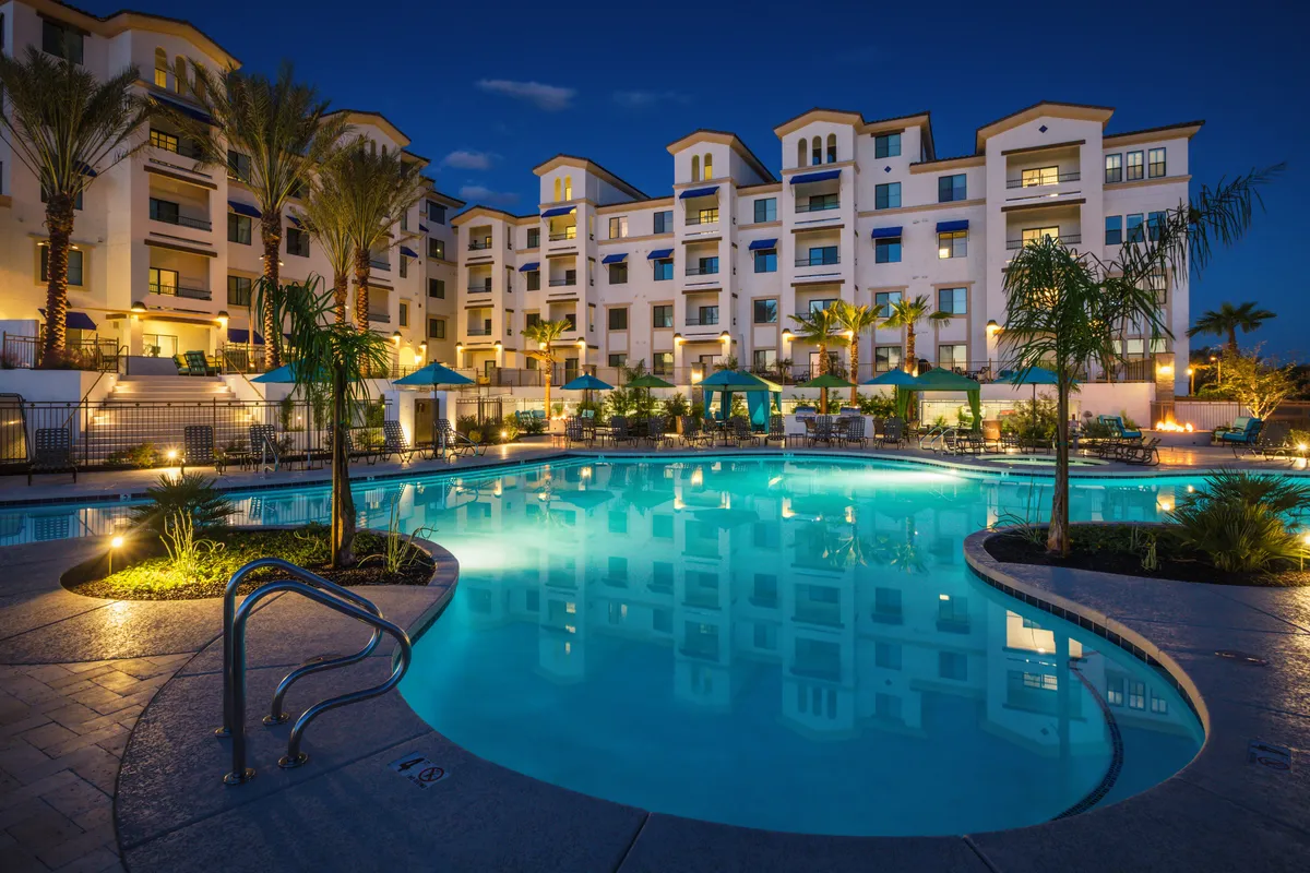 A luxurious pool area flanked by palm trees, featuring stylish lounge chairs and cabana tents. The pool is illuminated against the evening backdrop, creating an atmosphere of indulgence and comfort. Small groups of people can be seen relaxing and socializing around the pool, suggesting vibrant community life.