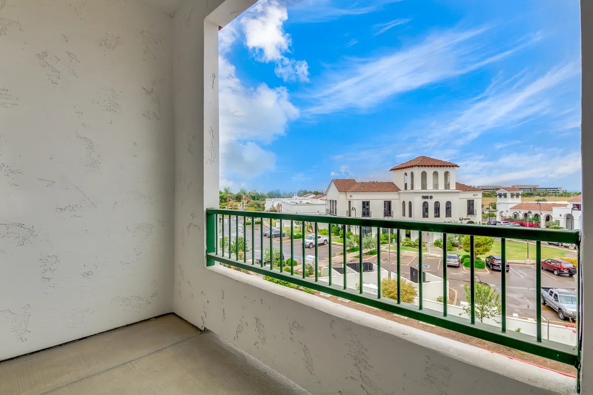 A tranquil exterior view of the building across the street, set against a blue sky with clouds. The green balcony railing adds a lively touch, and the scene captures the calm of the neighborhood with hints of daily life, such as people enjoying a morning coffee, reading a book, or admiring the view from their balconies.