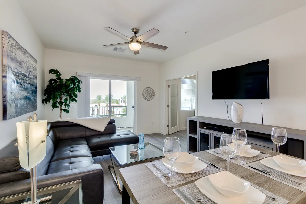 A spacious interior scene with a sofa, dining table, television, and a lush plant. The arrangement invites both relaxation and social interaction, with areas for shared meals, watching movies, and vibrant conversation. Bright natural light highlights the contemporary, comfortable furnishings.