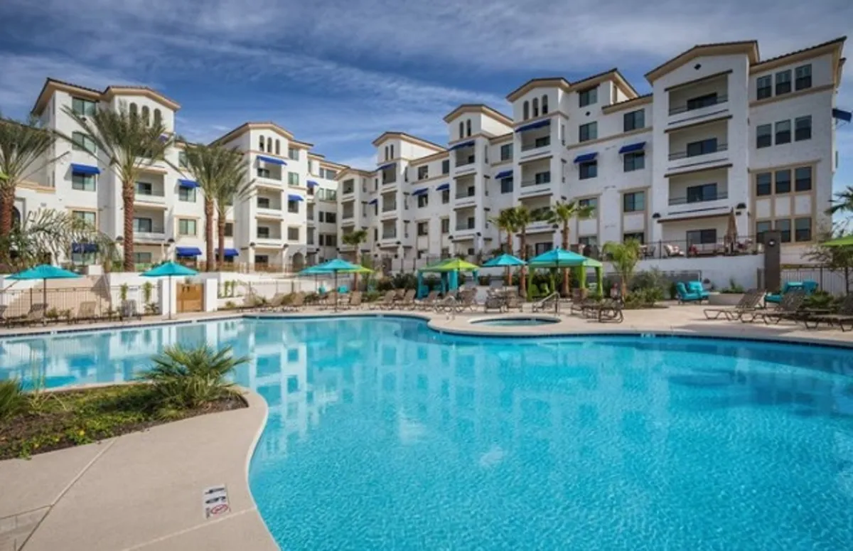 An inviting scene of a resort-style swimming pool surrounded by palm trees, blue umbrellas, and outdoor lounge chairs, with the apartment building in the background. The mood is leisurely and refreshing, suggesting post-golf relaxation.