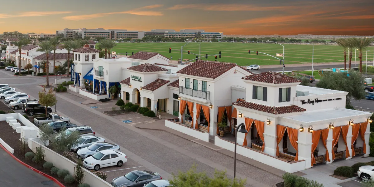 An exterior plaza scene with buildings featuring terracotta roofs, orange drapes adding a festive touch, and lush green landscaping. The area is designed for dining outdoors, shopping, and community events, with sunset lighting creating a warm and welcoming mood.