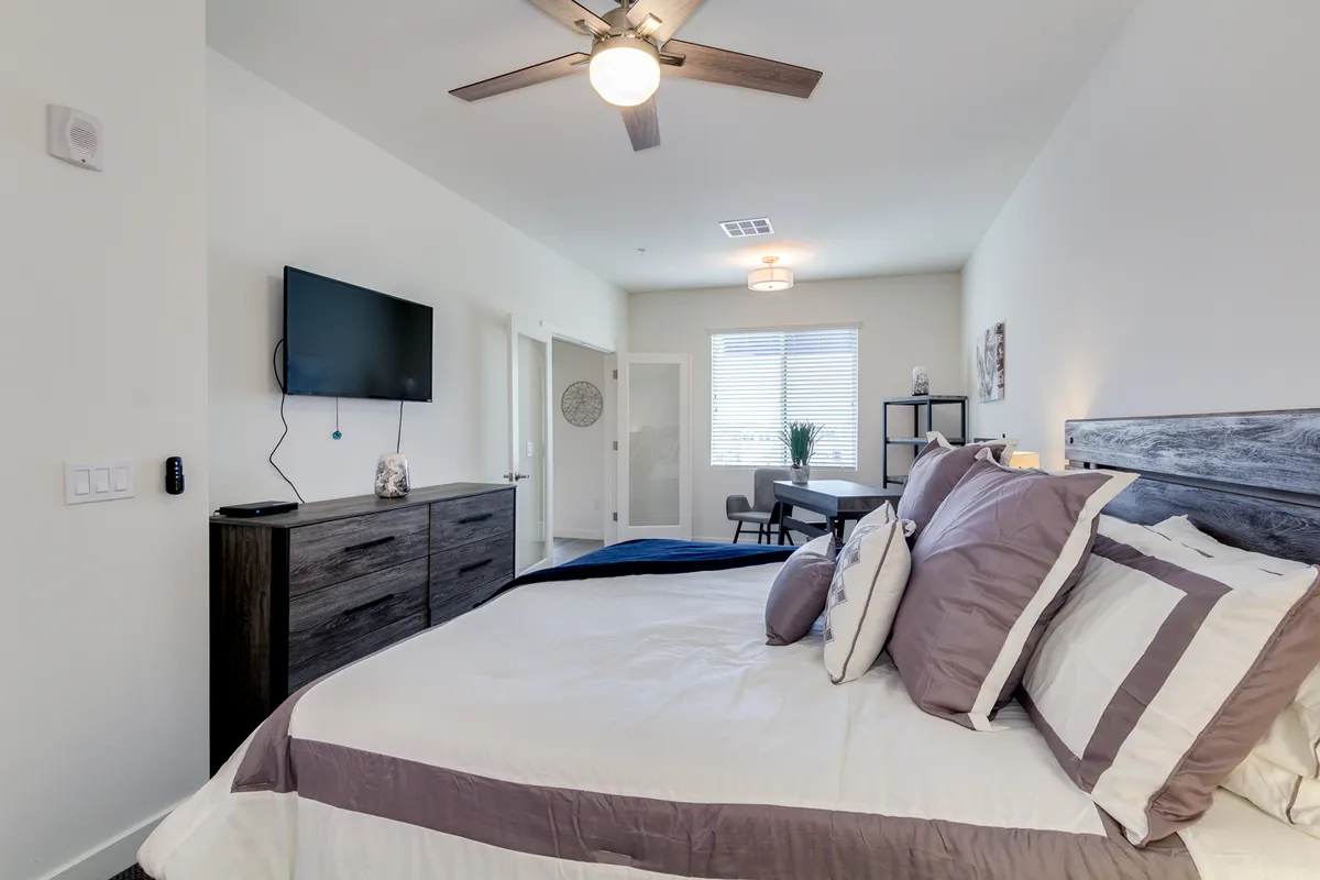 This image presents a cozy bedroom featuring a comfortable bed with decorative pillows, a wall-mounted TV, a dining table and chairs, and a window letting in natural light. The inviting layout makes it ideal for lounging, watching TV from bed, working at the desk, or enjoying the sunlight.