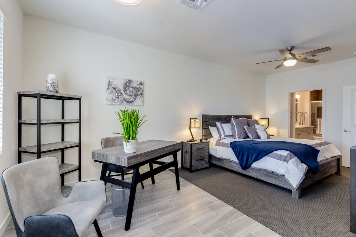 A serene room with a bed adorned with decorative pillows, a desk with a plant, and stylish shelves. The space feels cozy and inviting, emphasizing areas for both relaxation and productive work, with abundant natural light contributing to a tranquil atmosphere.