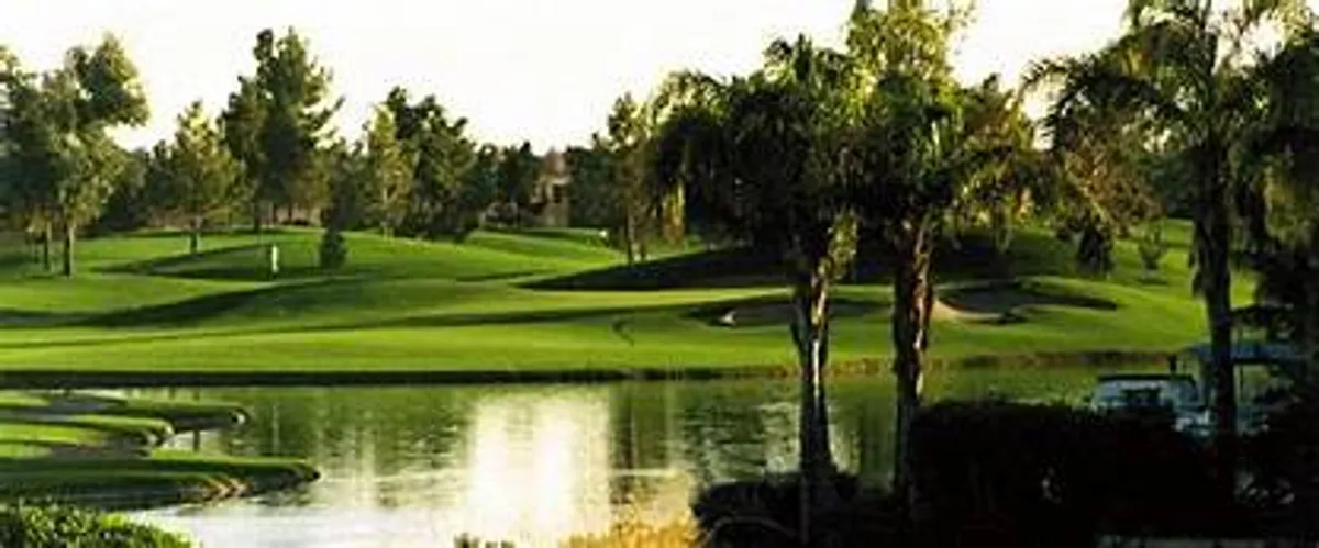 A sweeping exterior view of a lush green golf course, complete with calm water features, tall palm trees, and rolling hills under an open sky. The setting exudes tranquility and the appeal of the Arizona golf experience.